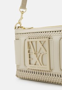 Beżowa torebka z tkaniny z gładkimi akcentami, złotymi elementami i tłoczoną plakietką z logo. Posiada konstrukcję o ustalonej formie oraz regulowany pasek.