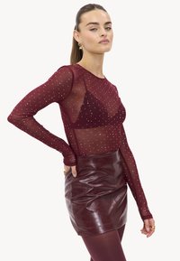 Bordeaux mesh lange mouw top met zilveren studs, gecombineerd met een glanzende bordeaux mini rok. Gestructureerde stof met doorschijnende designelementen.