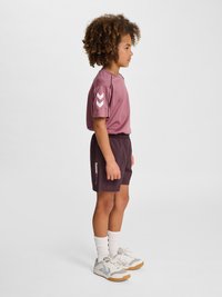 Barn med krøllet hår står sidelæns iført en lilla sportstrøje, mørke shorts, hvide sokker og grå sneakers på ensfarvet baggrund.