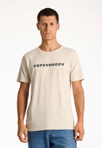 Camiseta de algodón beige con cuello redondo y mangas cortas; presenta "COPENHAGUE" impreso en negro en el pecho.