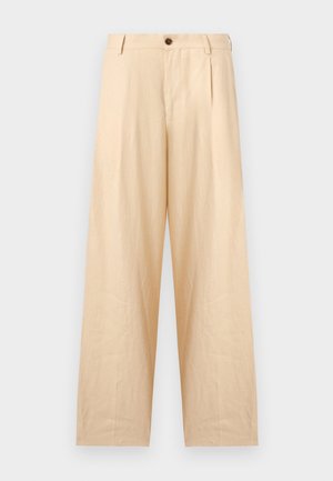 Mann mit lockigem dunklem Haar, der ein rostfarbenes kurzärmliges Hemd über einem weißen T-Shirt, beigen Hosen und schwarzen Sandalen trägt.