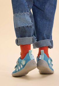 Scarpe da bambino blu con accenti bianchi a forma di triangolo, parte superiore testurizzata, suola in gomma e dettagli rossi giocosi. Indossate con denim arrotolato e calzini arancioni.