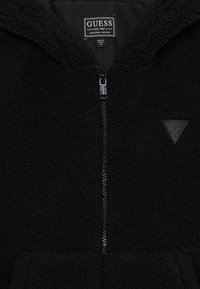 Juodas pliušinis zip-up džemperis su minkšta tekstūra, priekyje su užtrauktuku ir krūtinės logotipo pleistru. Apatinėje dalyje turi kengūros kišenę.