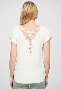Hvit topp med V-formet ryggdesign, intrikate blonderdetaljer, korte ermer og et strukturert stoff. Bakdelen har blomstermønster.