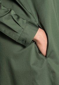 Grön, texturerad skjorta i tyg med knappad manschett och sidoficka. Materialet är slätt med en lätt glans, med rena linjer och en strukturerad design.