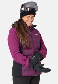 Lila isolierte Jacke mit schwarzen Akzenten, strukturiertem Stoff und Reißverschluss vorne. Schwarze Handschuhe. Große Skibrille und Stirnband.