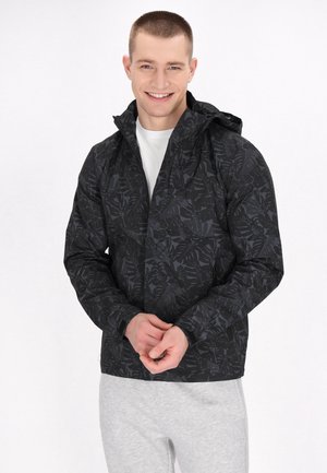 Zwarte jas met een volledig donker bladerenpatroon, met een capuchon en een rits aan de voorkant. Zit snugger, gecombineerd met lichtgrijze broek.