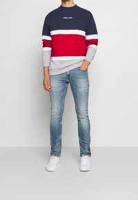Muž v námořnickém, bílém, červeném a šedém svetru Tommy Jeans, světlých modrých slim-fit džínách a bílých teniskách, stojící na jednobarevném pozadí.