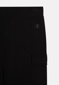Pantalons cargo noirs avec une texture lisse, dotés d'une poche latérale et d'un logo discret. Le tissu semble doux et extensible.