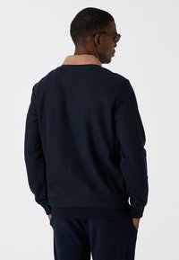 Granatowy sweatshirt z beżowym kołnierzem, ściągaczami przy mankietach i u dołu. Tkanina ma teksturowane wykończenie, a projekt charakteryzuje się luźnym krojem.