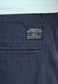 Primer plano del bolsillo trasero de unos pantalones azul marino con una etiqueta blanca bordada que dice "PETROL IND & Co." y detalles cosidos.