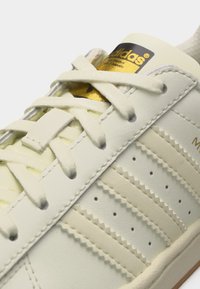 Gros plan sur une basket Adidas blanche avec des bandes crème, des lacets blancs et une étiquette Adidas dorée sur la languette.