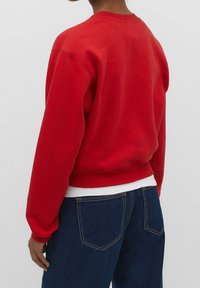 Personne portant un sweat-shirt rouge à manches longues sur une chemise blanche et un jean en denim bleu, montré de dos sur un fond uni.