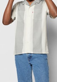 Homme portant une chemise à manches courtes à rayures verticales beige et grises avec poches poitrine et un jean bleu, sur un fond uni.