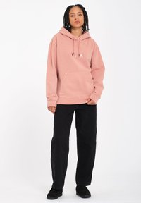 Hooded sweatshirt in soft pink with a kangaroo pocket and front logo. Paired with black loose-fitting pants and black shoes.
