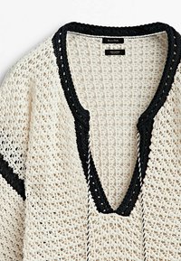 Pull en tricot au crochet de couleur crème avec des accents noirs. Il présente un col en V large, un tissu texturé et des détails en corde tressée au niveau de l'encolure.