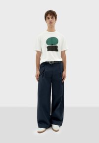 T-shirt bianca con grafica verde e nera, abbinata a pantaloni larghi blu navy. Calzature con suola bianca e marrone. Design semplice.