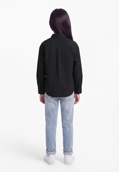 Pitkähiuksinen lapsi, jolla on musta pitkähihainen paita, vaaleansiniset farkut ja valkoiset lenkkarit, seisoo selin valkoista taustaa vasten.