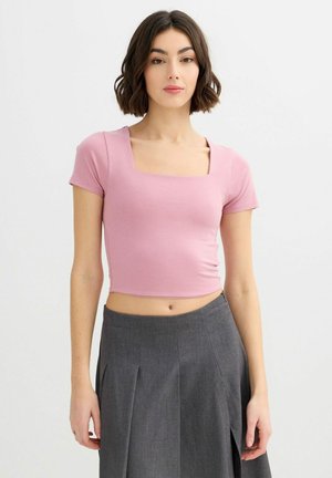 Mujer con cabello corto y castaño, vestida con un top corto rosa de escote cuadrado y una falda plisada gris de talle alto, frente a un fondo liso.