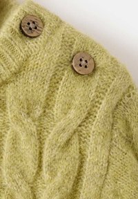 Stickad tröja i ljusgrön med ett kabelmönster och två bruna knappar vid halsringningen. Texturerad, mysig tyg.