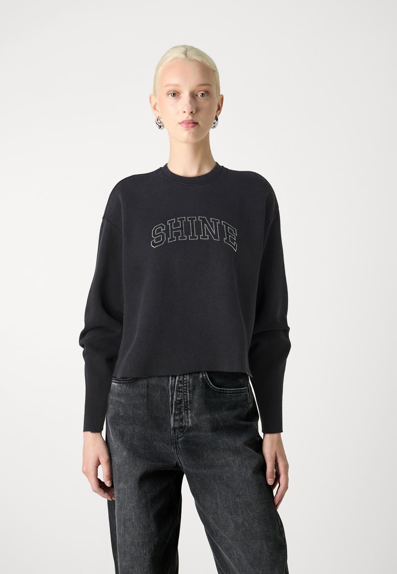 Sweatshirt noir court avec "SHINE" en couture contrastante, doté de manches larges et d'un col rond, associé à un jean noir.