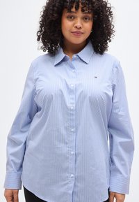 Ανοιχτό μπλε και λευκό ριγέ πουκάμισο με κουμπιά, μακριά μανίκια και μυτερό γιακά, με μικρό λογότυπο στην αριστερή περιοχή του στήθους.