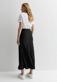 Jupe maxi noire et soyeuse avec une texture douce, associée à un t-shirt blanc ajusté et des baskets blanches. Vue par l'arrière.