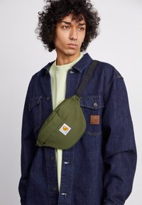 Borsa a tracolla verde in tessuto indossata sopra una giacca di jeans. La borsa presenta un design curvo con una toppa con il logo. Lo sfondo è neutro.