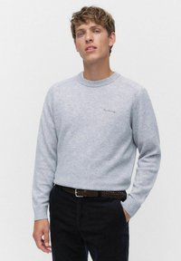 Ljust grått stickat tröja med rund halsringning, ribbade ärmslut och en liten broderad logotyp på bröstet. Bärs med mörka byxor och ett flätat bälte.