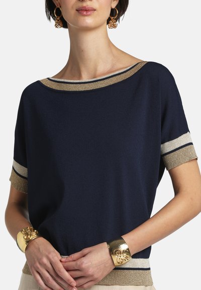 Frau trägt ein marineblaues Kurzarmoberteil mit gold- und beigen Verzierungen, große goldene Creolen und passende auffällige goldene Manschettenarmbänder.