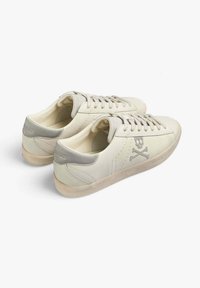 Sneaker in pelle beige con accenti grigi, leggermente texturizzati, con punta tondeggiante, lacci piatti e un sottile logo con teschio e ossa incrociate.