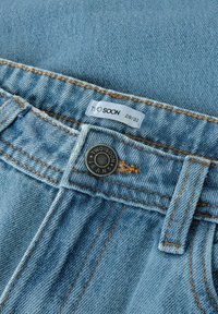 Svetlo modre kavbojke imajo črn gumb z izgraviranim napisom "TWO SOON JEANS" in oznako za velikost 28/32. Vidne podrobnosti šivanja.
