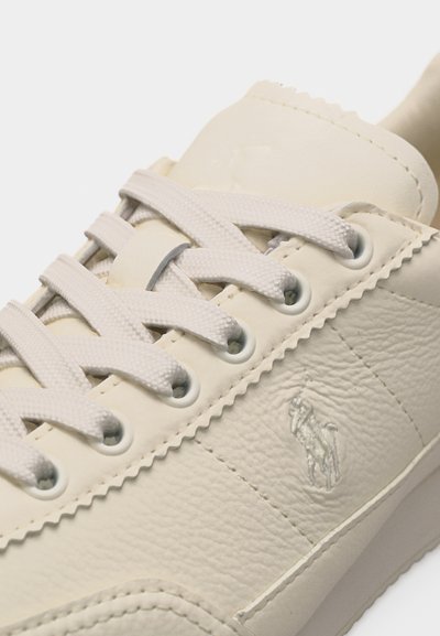 Κοντινό πλάνο μπεζ δερμάτινου αθλητικού παπουτσιού με κορδόνια στο ίδιο χρώμα και ανάγλυφο λογότυπο με παίκτη πόλο πάνω σε άλογο στην πλαϊνή πλευρά.