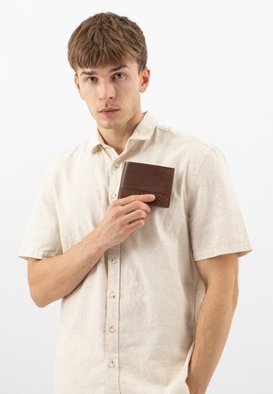 Junger Mann in beige Hemd hält eine braune Lederbrieftasche dicht an die Brust, blickt nach vorne vor schlichtem weißem Hintergrund.