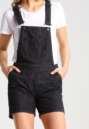 Osoba ubrana w czarne krótkie ogrodniczki z denimu z przednią kieszenią i białą koszulkę z krótkim rękawem, ręce w bocznych kieszeniach.