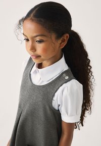 Graues ärmelloses Kleid mit Falten, über einer weißen Bluse mit Kragen getragen. Verfügt über runde Knöpfe an der Schulter und eine weiche Stofftextur.