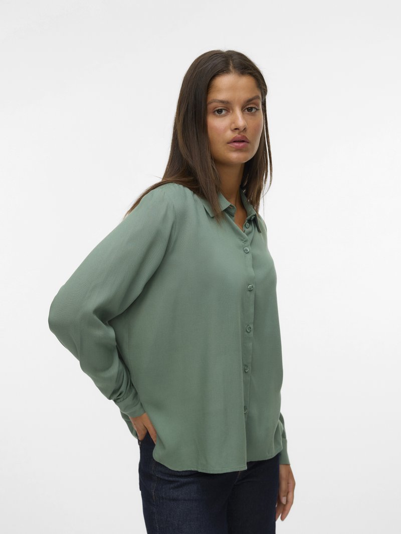 Chemise boutonnée en tissu vert doux et texturé, avec un col, des manches longues et une coupe décontractée. Associée à un jean foncé.
