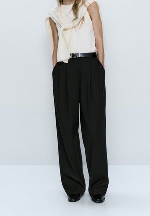 Black pleated trousers with a high waist, a white sleeveless top, and a light-colored sweater draped over shoulders; paired with black shoes.
