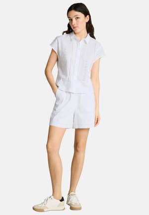 Frau trägt ein weißes, kurzärmliges Hemd mit Knöpfen und passende Shorts mit Lochstickerei, beige-weiße Turnschuhe, steht mit den Händen in den Taschen.