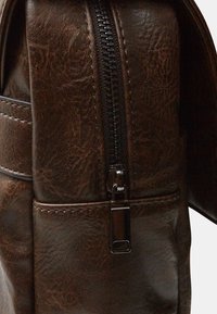 Borsa in pelle marrone con texture, caratterizzata da una zip prominente e hardware elegante, con dettagli di cucitura lungo i bordi e tasche laterali.