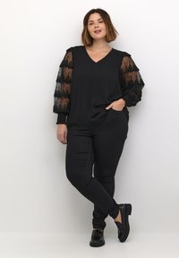 Černý top s průhlednými strukturovanými rukávy s vrstvenými detaily; doplněno černými slim kalhotami a lesklými černými botami.