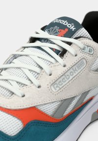 Zbliżenie na białe, niebieskie i pomarańczowe sneakersy Reebok z białymi sznurowadłami, siateczkowym materiałem, zamszowymi detalami oraz logo marki na języku i boku.