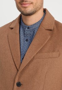 Cappotto di lana marrone con rever a lancia, chiusura con un bottone singolo e taschino sul petto, indossato sopra una camicia blu con motivo e bottoni.