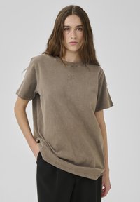 Junge Frau mit langen braunen Haaren trägt ein lockeres braunes Kurzarmhemd und schwarze Hose, steht vor einem schlichten hellgrauen Hintergrund.