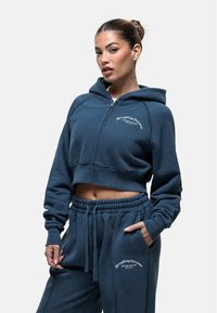 Korte blauwe hoodie met een rits, voorzien van een voorkantzak en geborduurde tekst. Gecombineerd met bijpassende joggingbroek met trekkoord.