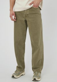 Pantalons en coton vert olive avec une coupe droite, dotés de poches avant et d'une taille visible, associés à des baskets grises et beiges.