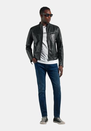 Uomo che indossa giacca di pelle nera, maglietta bianca, jeans blu, scarpe sportive nere e occhiali da sole, in piedi con una mano in tasca.