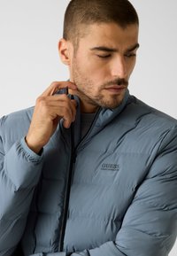 Jasnoniebieska pikowana kurtka z suwakiem z przodu, pikowaną teksturą i czarnym logo. Posiada stojący kołnierz i elastyczne mankiety.