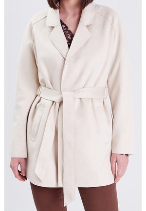 Beige belted coat with lapel collar worn over brown patterned top and brown pants, shown from neck to mid-thigh.