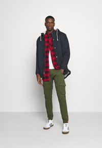 Svart jacka med huva, röd- och svartrutigt skjorta, vit t-shirt, olivgröna cargobyxor, vita sneakers med bruna accenter.